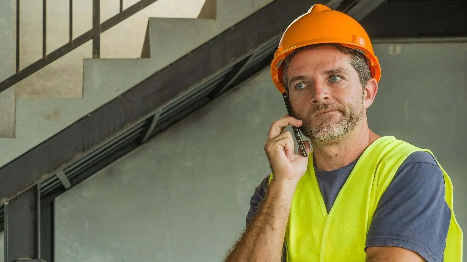 Ein Handwerker mi einem Schutzhelm und einer gelben Warnweste hält sich das Smartphone ans Ohr und guckt nachdenklich.
